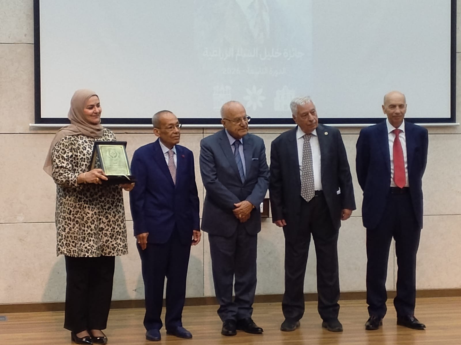 WhatsApp Image 2026-08-29 at 43332 PM1 - عروبة الاخباري | Oroba News Woman in a leopard print dress and hijab holds a plaque during an award ceremony on a stage with four suited men observing.