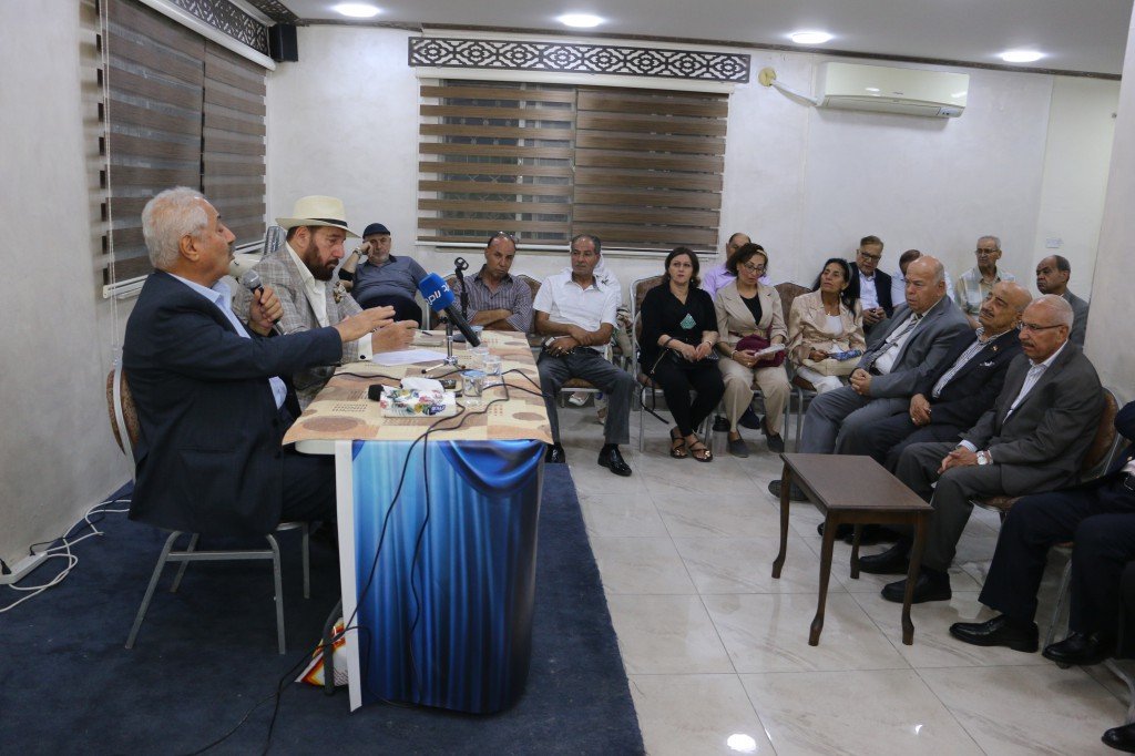 WhatsApp Image 2026-08-31 at 122508 AM - عروبة الاخباري | Oroba News A man in a suit speaks into a microphone at a panel table while another man in a hat sits beside him; audience listens in a small conference room.