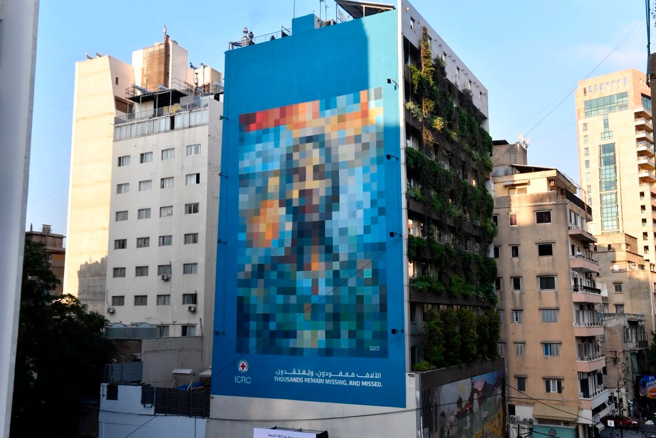 Blue billboard on a high-rise building featuring a pixelated mural about missing people, with ICRC logo and the phrase 'Thousands remain missing, and missed.' in English and Arabic