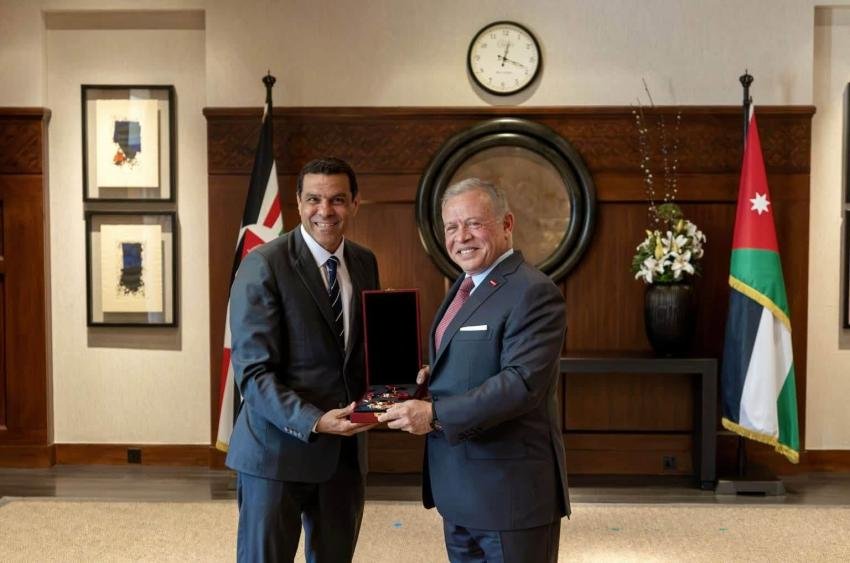 Two businessmen in suits exchange a red presentation box with a medal in a formal room with flags and a clock behind them.