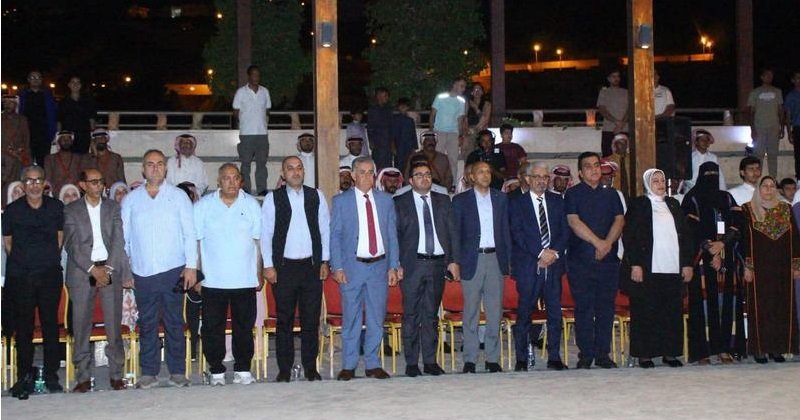 Line of formally dressed people standing in a row at an outdoor night ceremony, with spectators behind them.