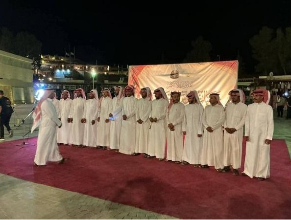A row of men in white traditional robes (kanduras) standing on a red carpet at an outdoor nighttime event, facing a speaker in the foreground with a banner backdrop behind them.