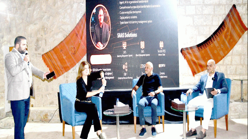 file2 - عروبة الاخباري | Oroba News Four people on a stage panel discussion; moderator stands left with a microphone, three panelists sit in blue chairs with a large banner behind them.