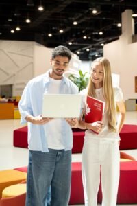 image1 - عروبة الاخباري | Oroba News Two students stand in a bright, modern lounge looking at a silver laptop together; the woman holds a red notebook labeled Canadian University Dubai.