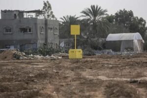 Desolate construction or conflict site with a damaged building, barbed wire, and a yellow warning sign on a post amid dirt and debris.