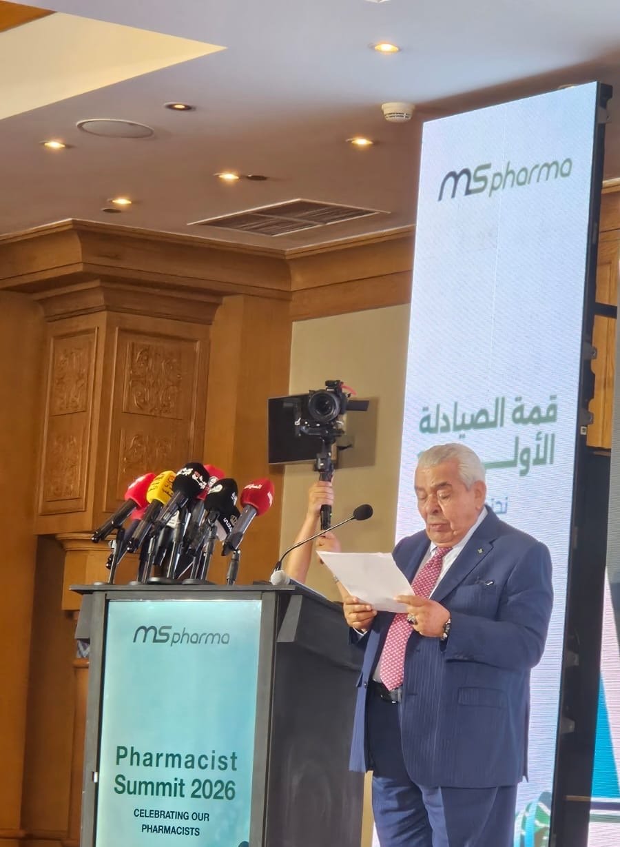 Man in a dark blue suit reading from a paper at a podium with multiple microphones during the Pharmacist Summit 2026, MSpharma backdrop behind him.