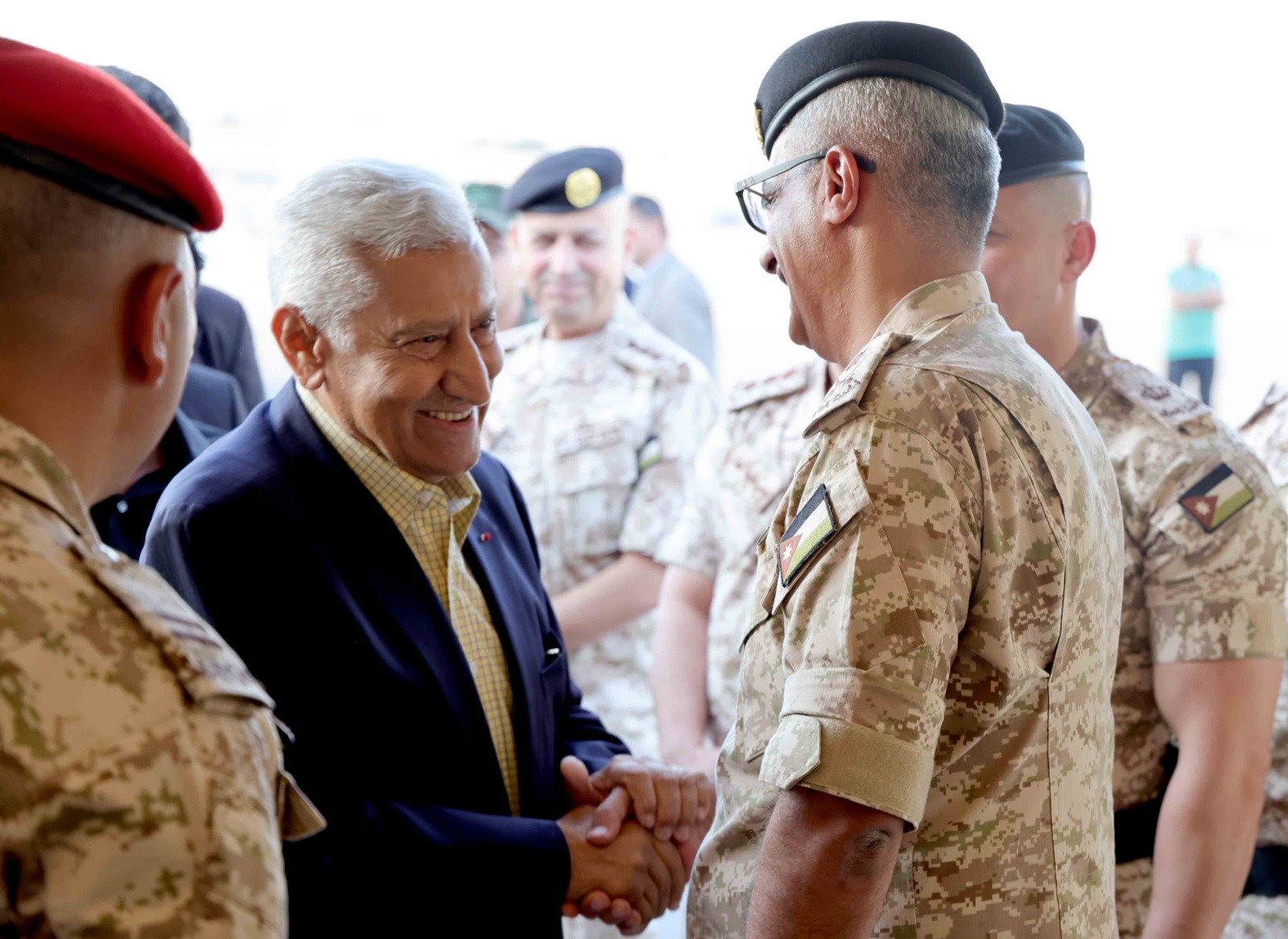 Civilian man in a blazer greets a camouflaged soldier with a handshake among a group of uniformed personnel, smiling faces visible in the background.