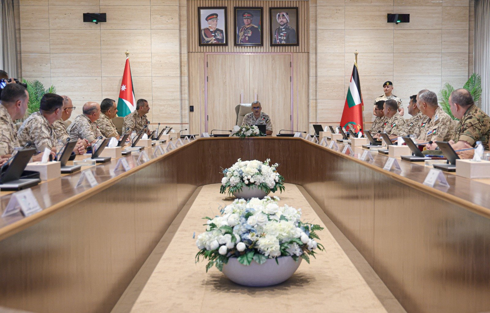Military officers sit around a U-shaped conference table in a formal meeting room, with Jordan and Kuwait flags flanking the head table and white floral arrangements in the foreground.