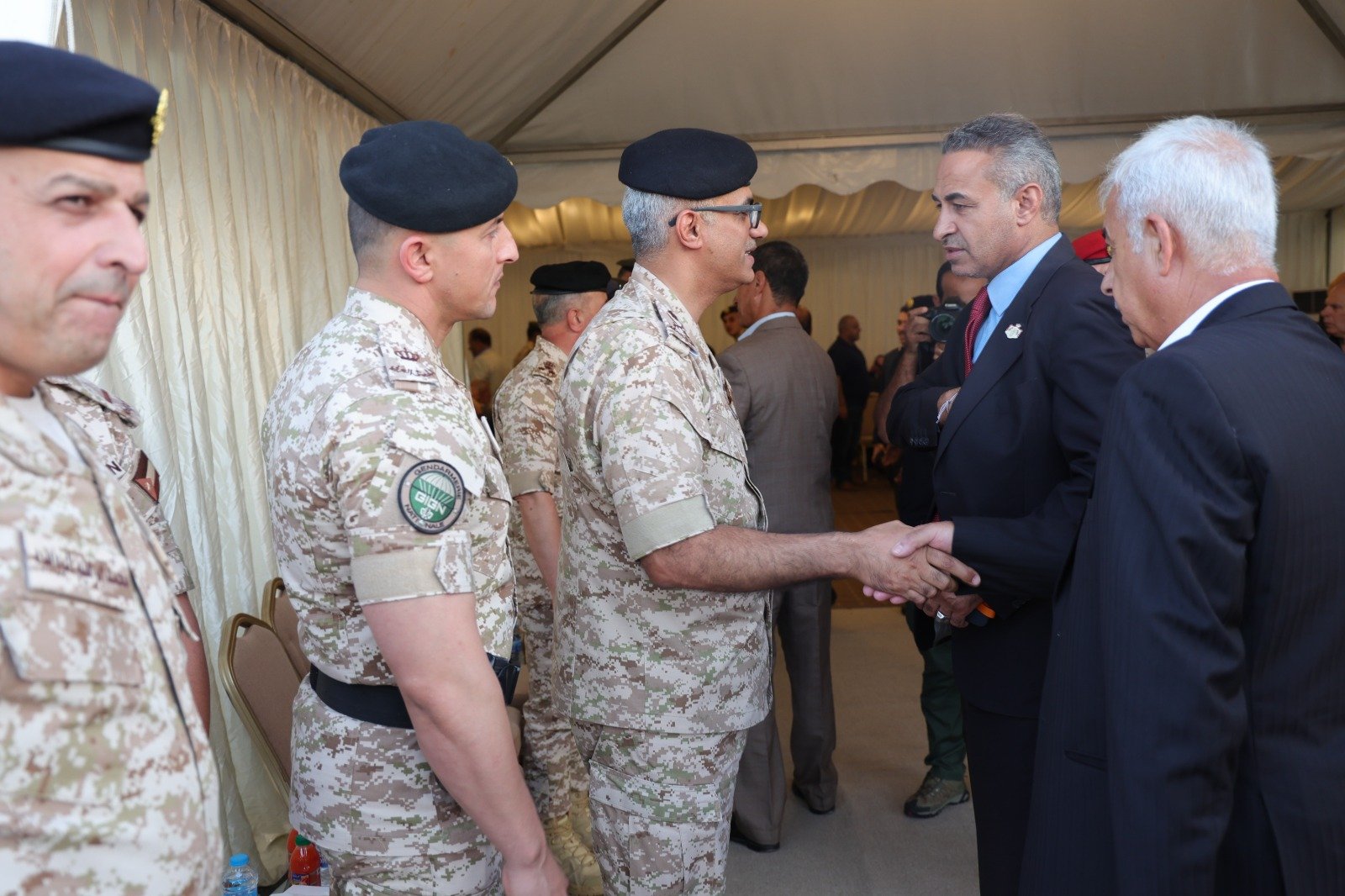 Military officers in camouflage shake hands with suited officials at a formal event inside a tent.