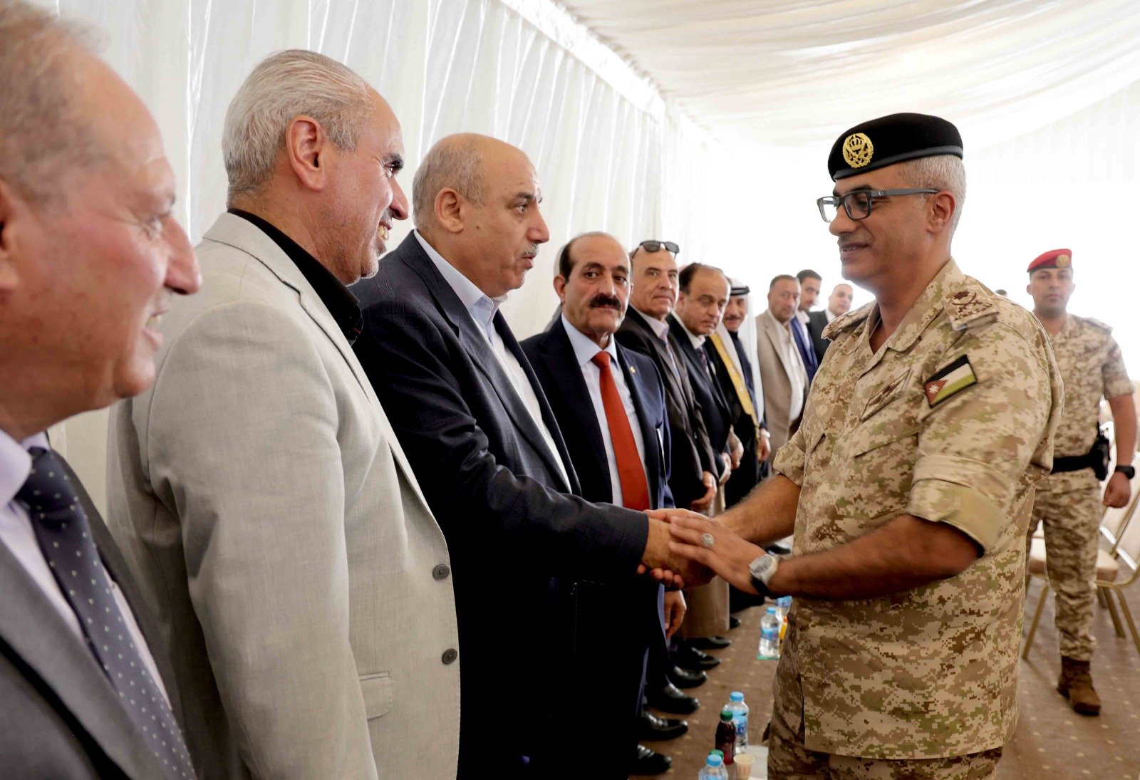 Military officer in desert camouflage shakes hands with a row of suited officials inside a white tent, as other uniformed personnel stand in the background.