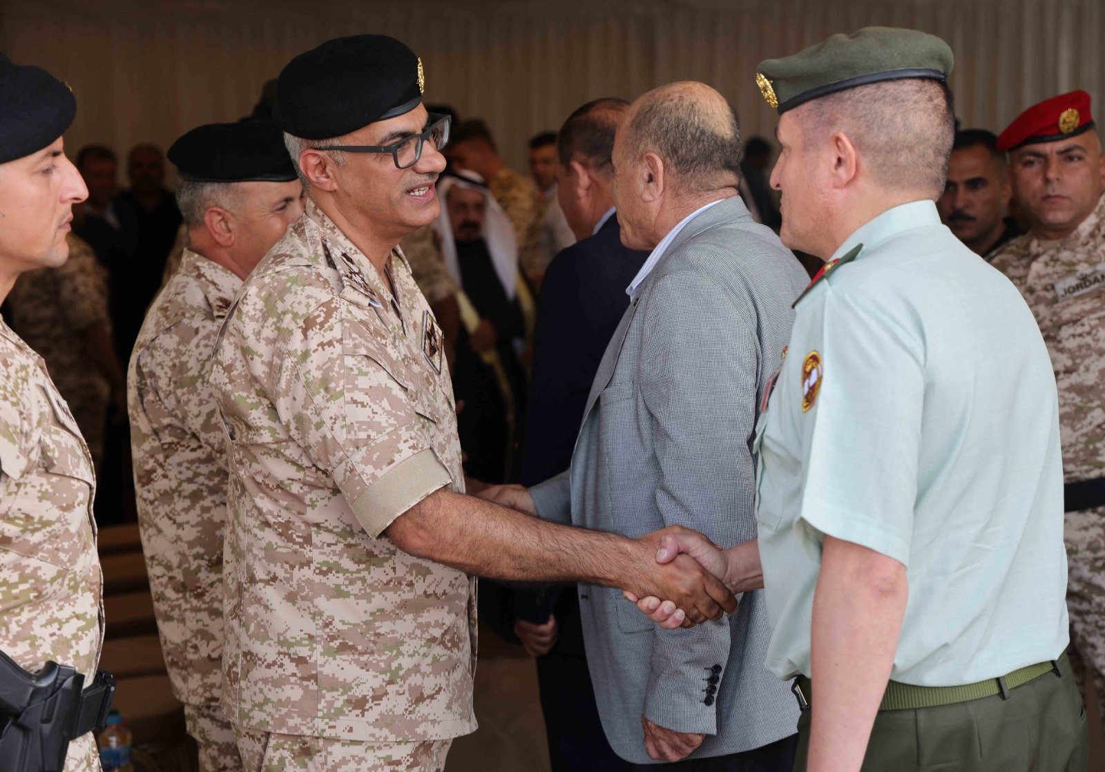 Military officer in camouflage shaking hands with a civilian man in a gray blazer at a formal event, other uniformed personnel in the background