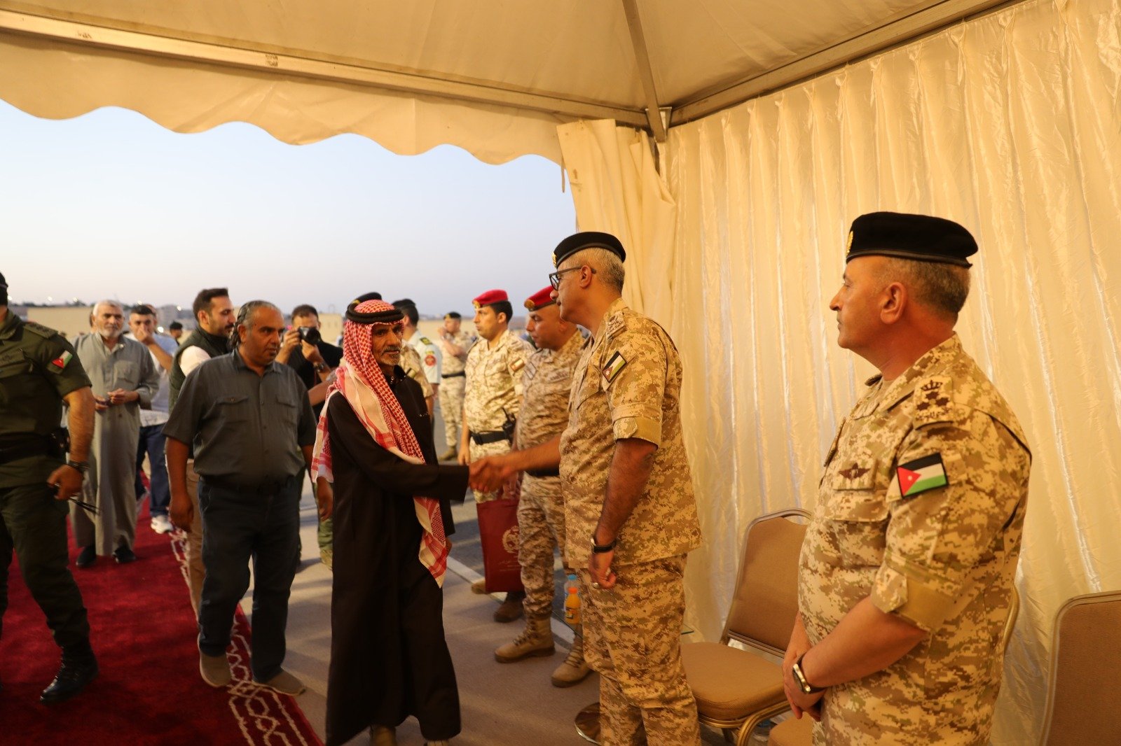 Military officers in desert camouflage shake hands with a man in traditional Arab attire under a tent.