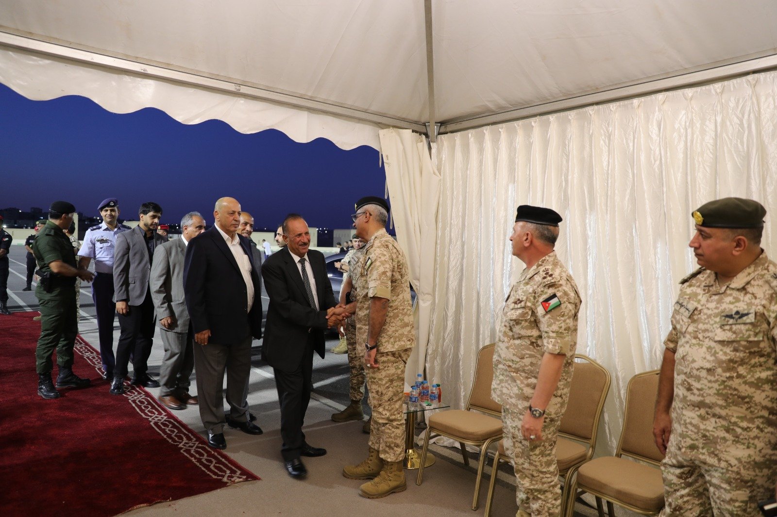 Officials in suits greeting a uniformed military officer with a handshake under a white tent at dusk on a tarmac ceremony.