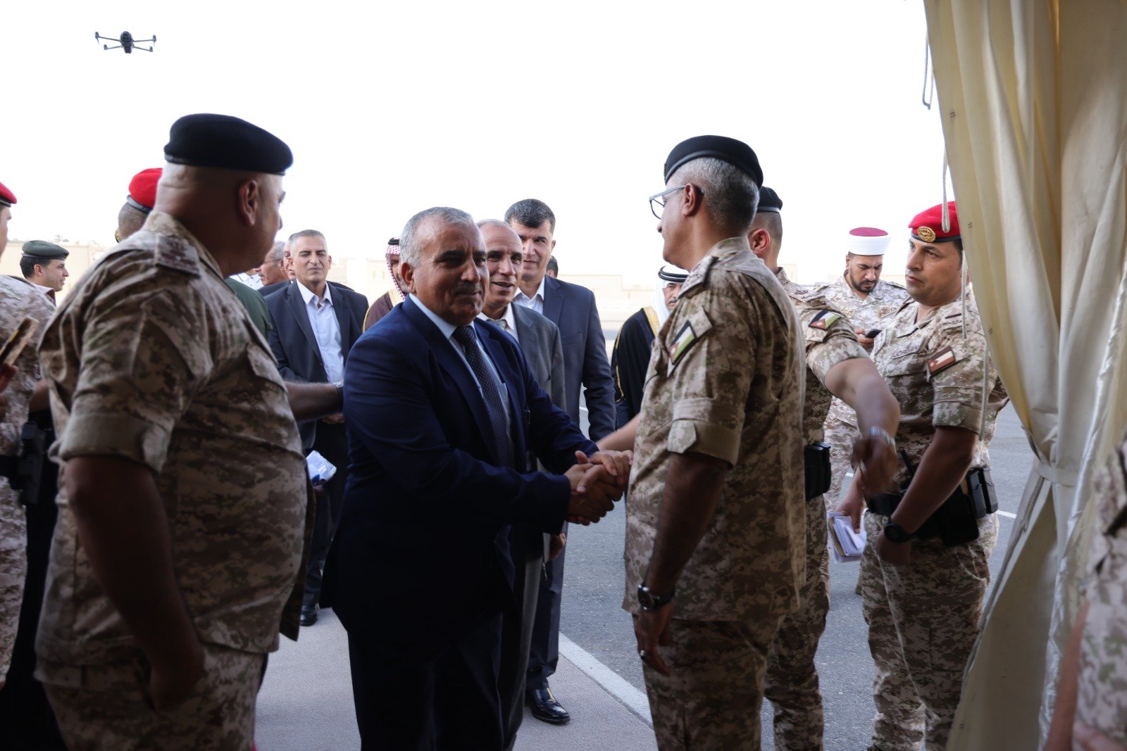 Dignitary in a dark suit shakes hands with a soldier in camouflage uniforms on a tarmac, with other officials watching nearby under a tent.