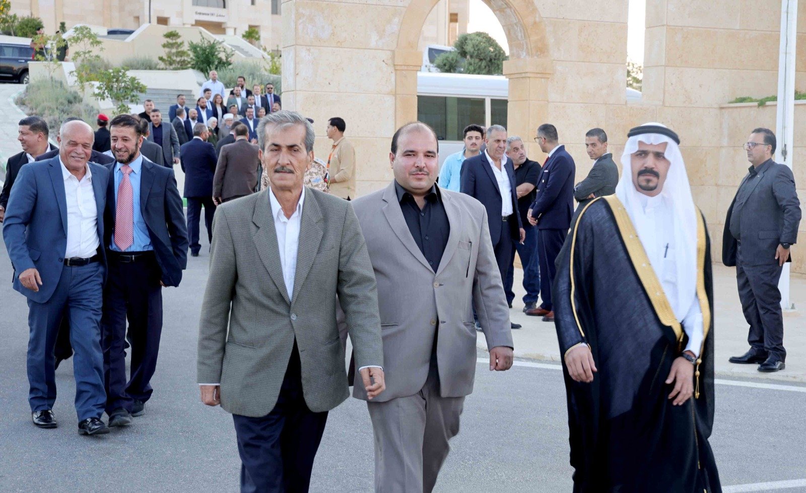 Group of men in suits walking outdoors past a beige stone arch, led by a man in a gray suit; a man in traditional robes stands to the right on the path.