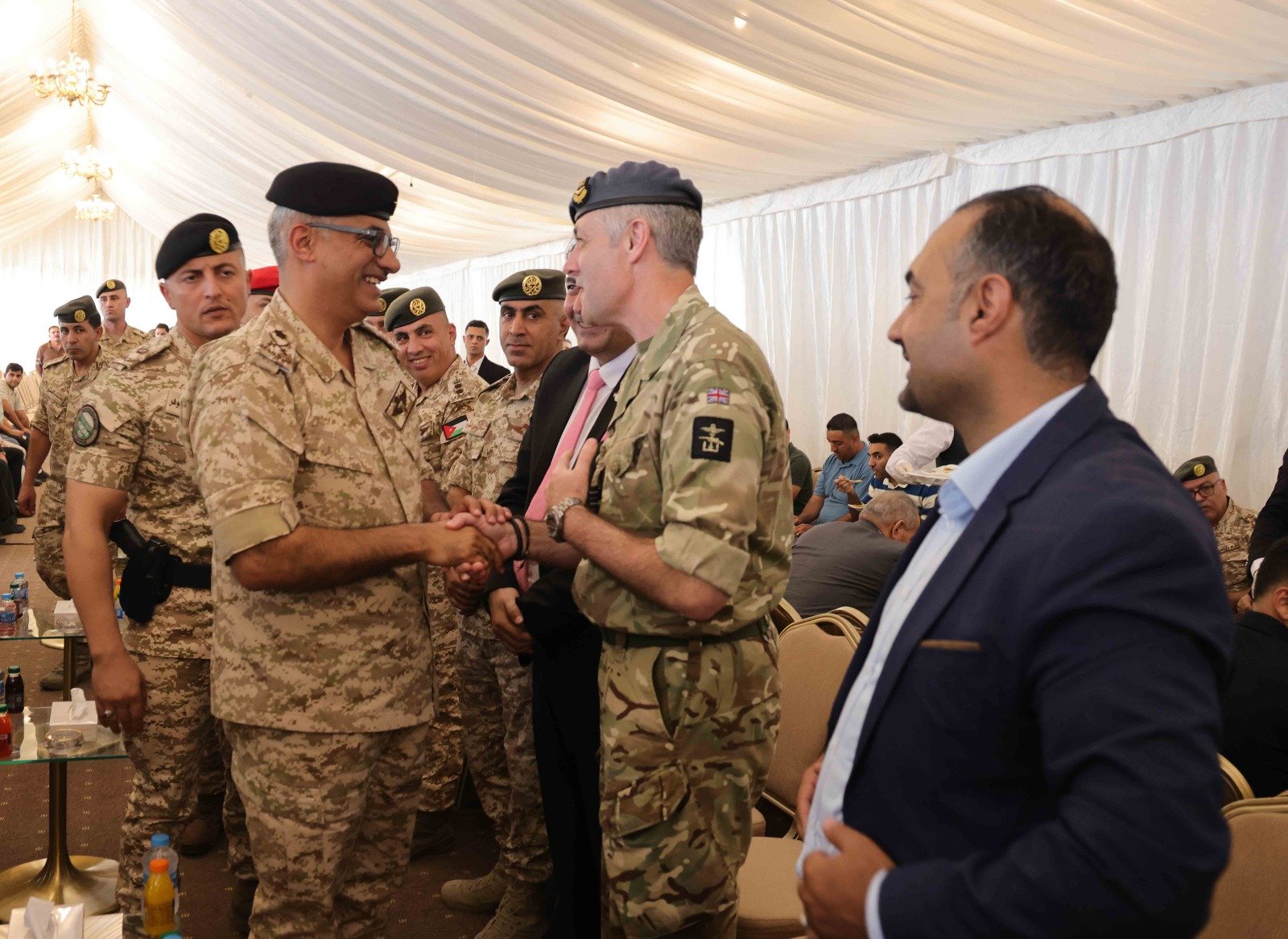 Soldiers in camouflage uniforms shake hands and smile at a formal indoor event under a white tent.