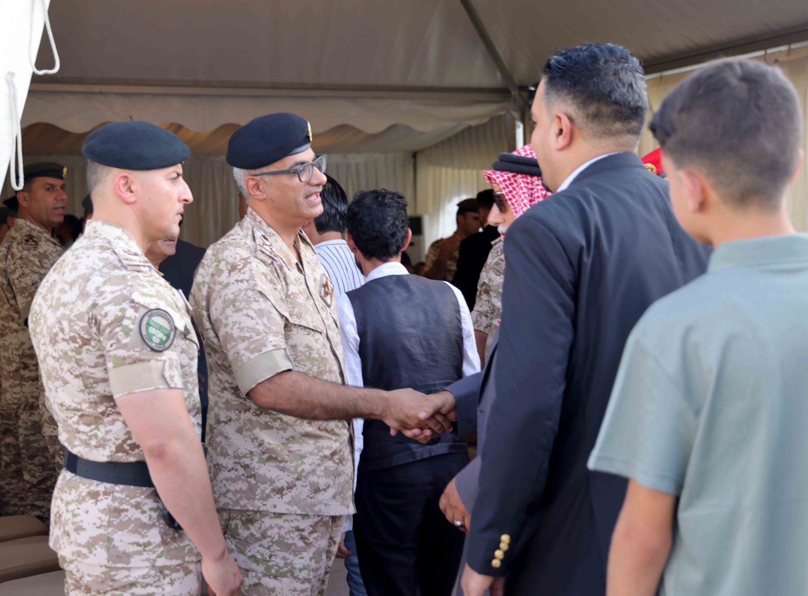Two soldiers in camouflage shake hands during a formal event under a tent, with others nearby.