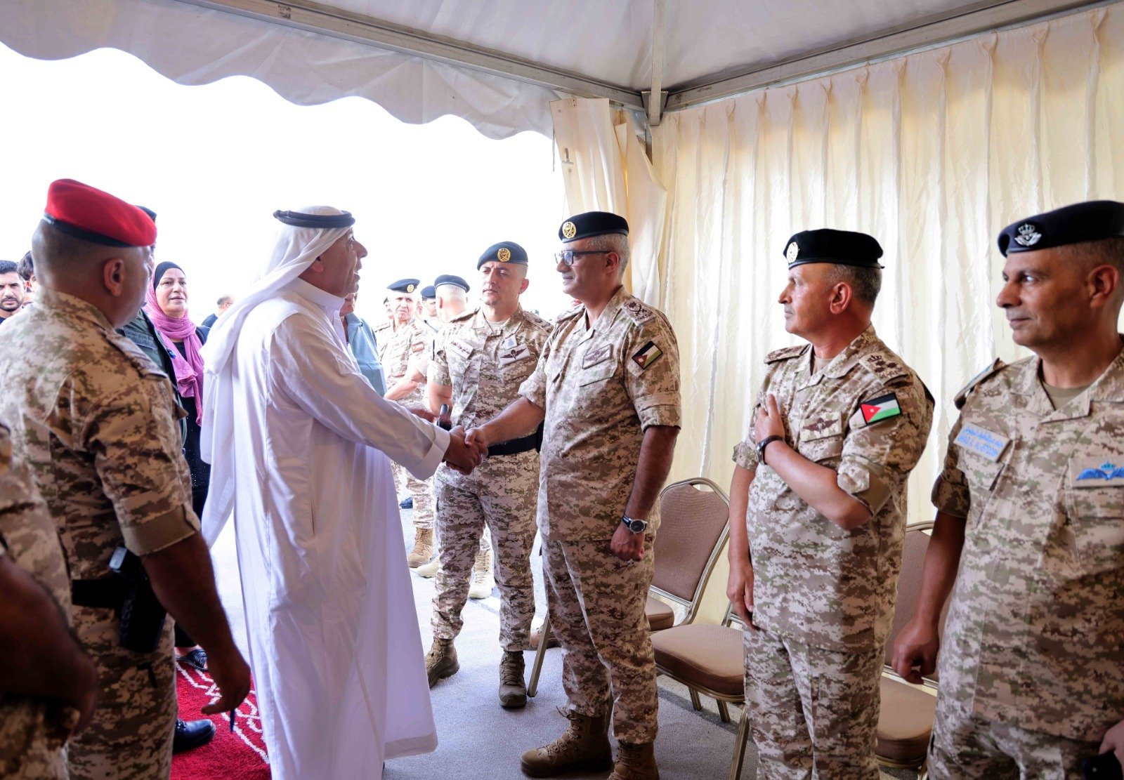 Man in a white thobe shakes hands with a Jordanian military officer as soldiers stand in a tent.