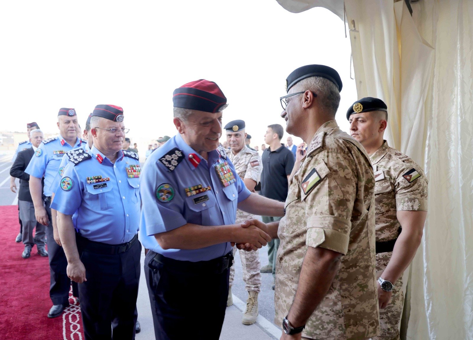 Senior officers in blue and camouflage uniforms shake hands on a red carpet at a formal military event outside a tent, medals on display