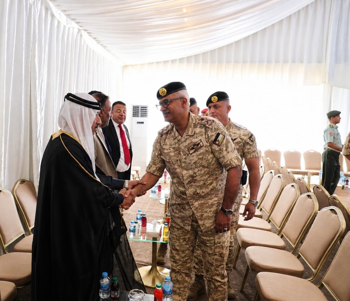 Senior military officer in desert camouflage shakes hands with a man in a black bisht and white headscarf inside a white ceremonial tent; chairs line the room.