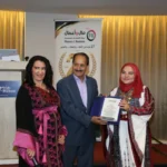Three people pose on stage as an award is handed to a woman in a red hijab, flanked by a man in a suit and another woman in a pink-black dress, at a conference.
