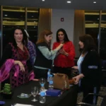 Group of women in a conference room stand around a black-clothed table with water bottles and a brown handbag on it, chatting as a banner in Arabic stands to the left.
