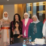 Group of seven women and a girl posing for a photo at a formal event, wearing colorful traditional dresses and hijabs, around a table with drinks.