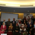 Group photo at an awards or recognition event with women in hijabs and men in formal attire in a conference room, some holding plaques.