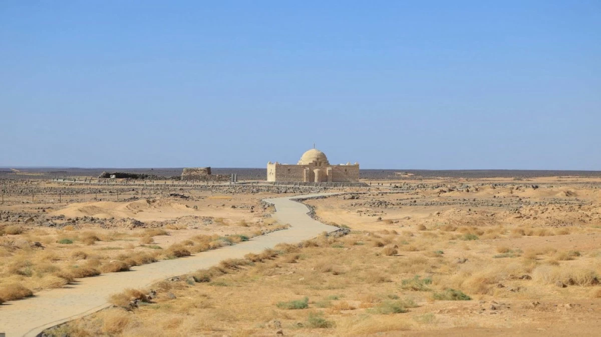 Desert landscape with a winding path leading to a small domed fortress on the horizon under a blue sky.