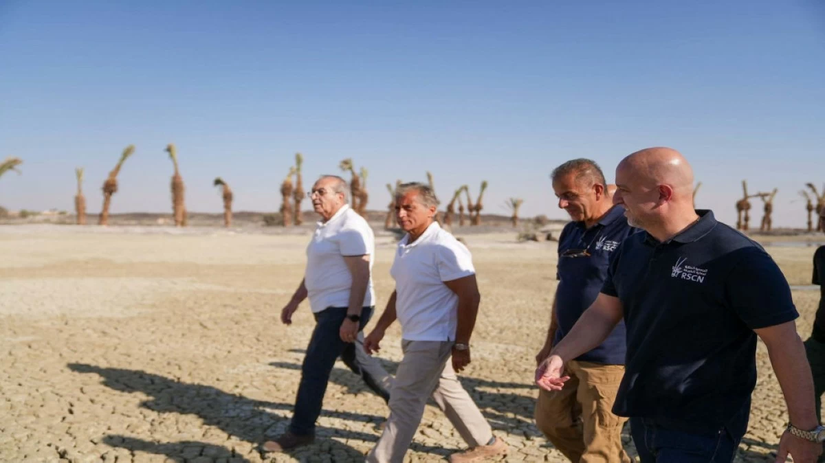 Four men walk across a cracked, arid desert landscape under a clear blue sky, with palm trees visible in the background.