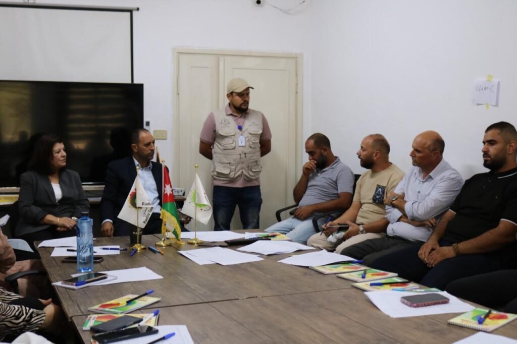People sit around a conference table with papers and notebooks; a man in a beige vest stands at the doorway addressing the group.