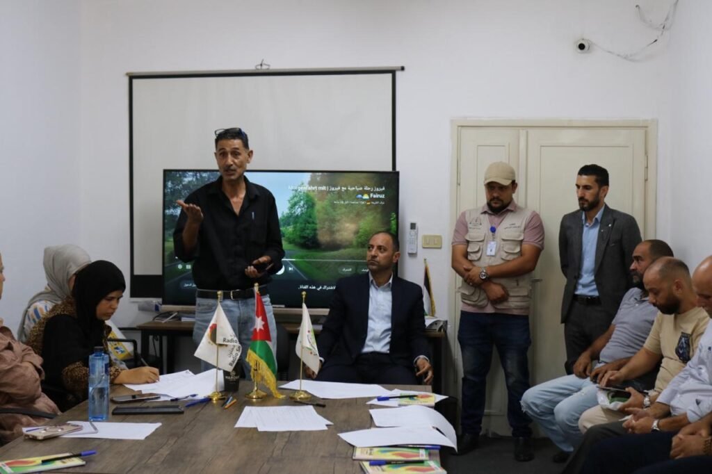 Man in a black shirt speaks while gesturing at a meeting room, with attendees seated around a table and a TV screen showing a nature scene behind him. Flags sit on the table in front of the group.