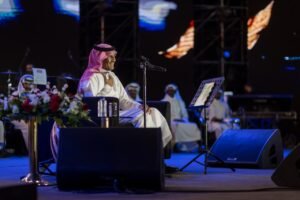 Man in traditional white thobe and red-and-white keffiyeh sits on a stage with a microphone, flower arrangement nearby, ready to speak.