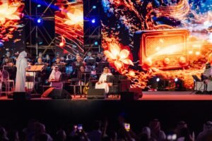 Orchestra on stage with a conductor in white robes, musicians reading from stands, and a vibrant digital backdrop; audience films the performance.