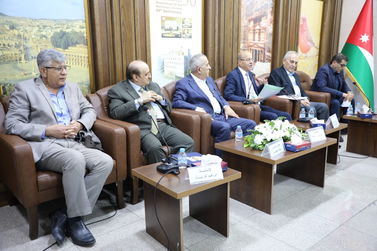 Six men in suits sit on brown leather chairs at a panel discussion, with microphones and nameplates on small tables.