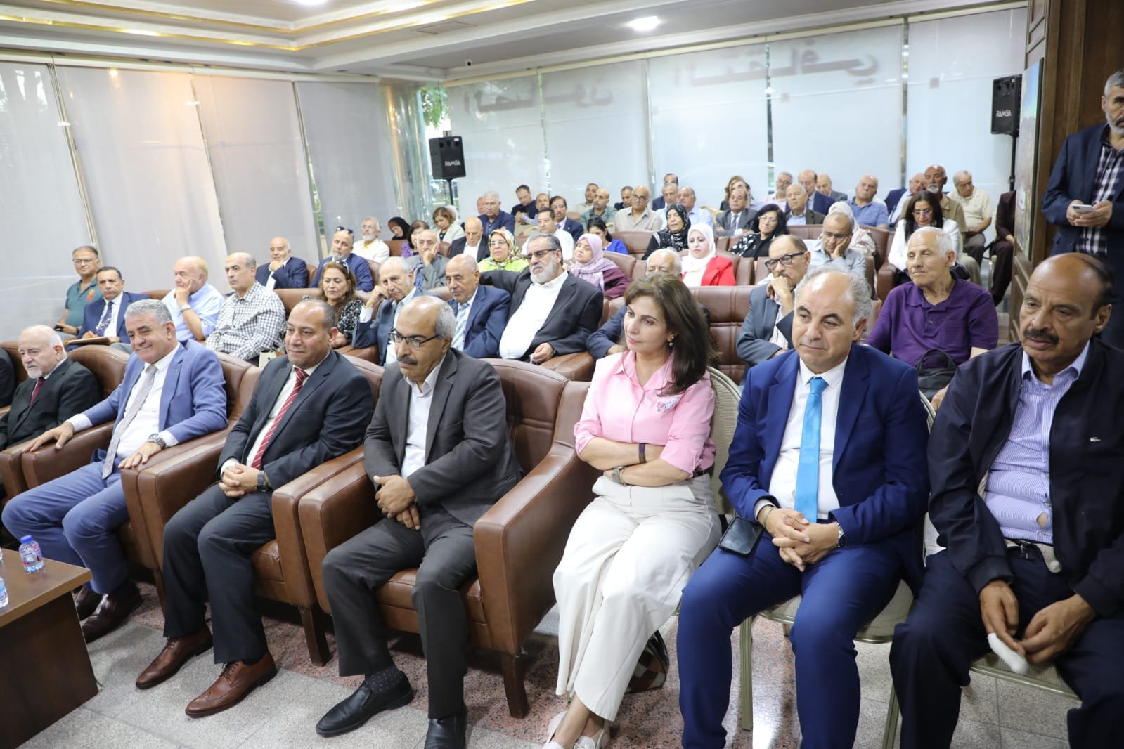 Audience seated in leather chairs at a formal meeting, listening to a presenter in a bright conference room.