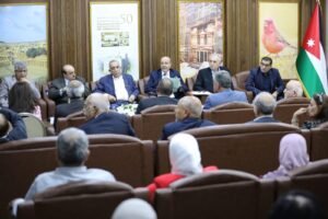 Formal panel meeting with officials seated in a semicircle, one man speaking into a microphone while others listen; Jordan flag visible on the right.