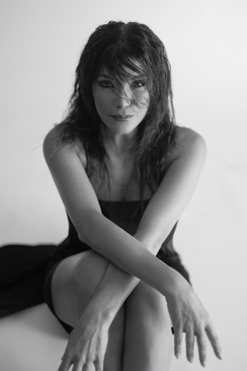 WhatsApp Image 2026-09-02 at 73342 PM3 - عروبة الاخباري | Oroba News Black-and-white portrait of a woman seated with her legs crossed, arms resting on her knees, looking softly at the camera.
