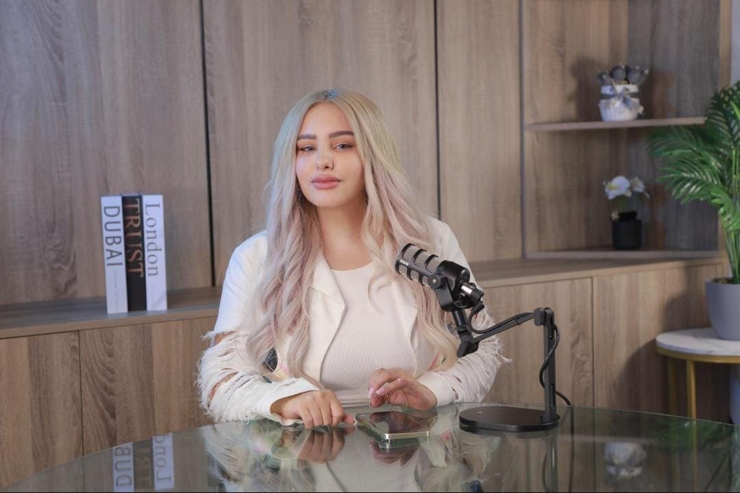 Woman with long blonde hair sits at a glass table in a podcast studio, microphone ready in front of her, shelves and plants in the background.