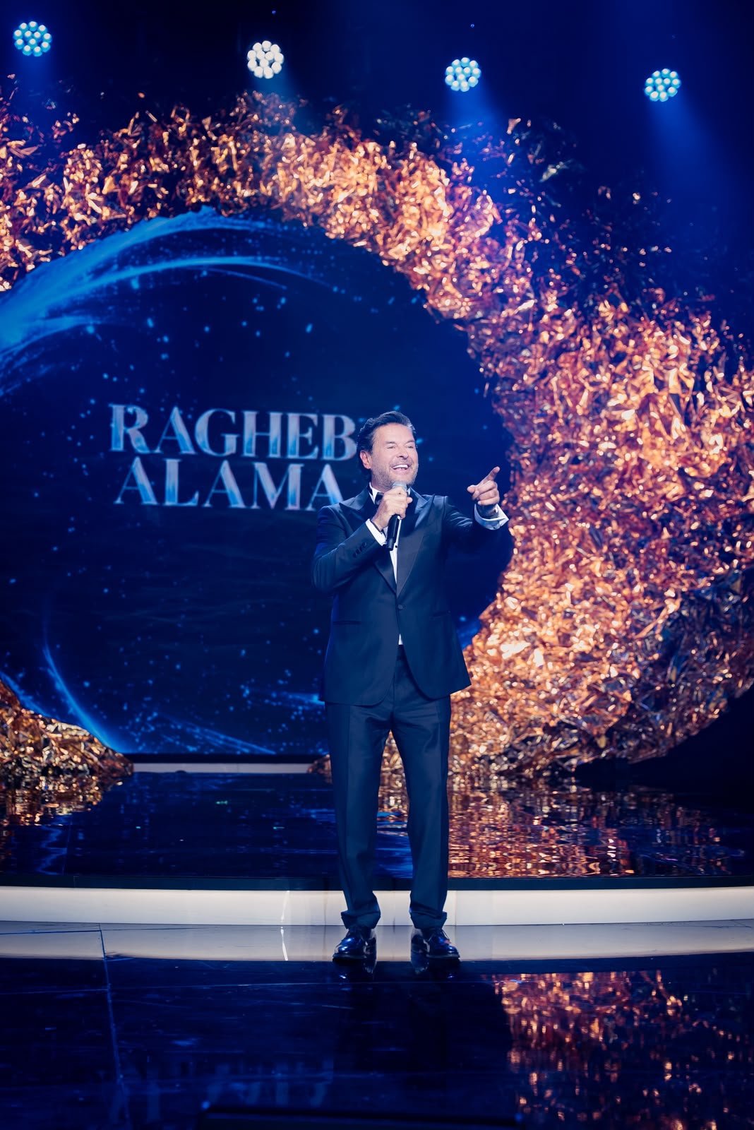 Male singer in a tuxedo performs on a stage with blue lights and a gold confetti backdrop.