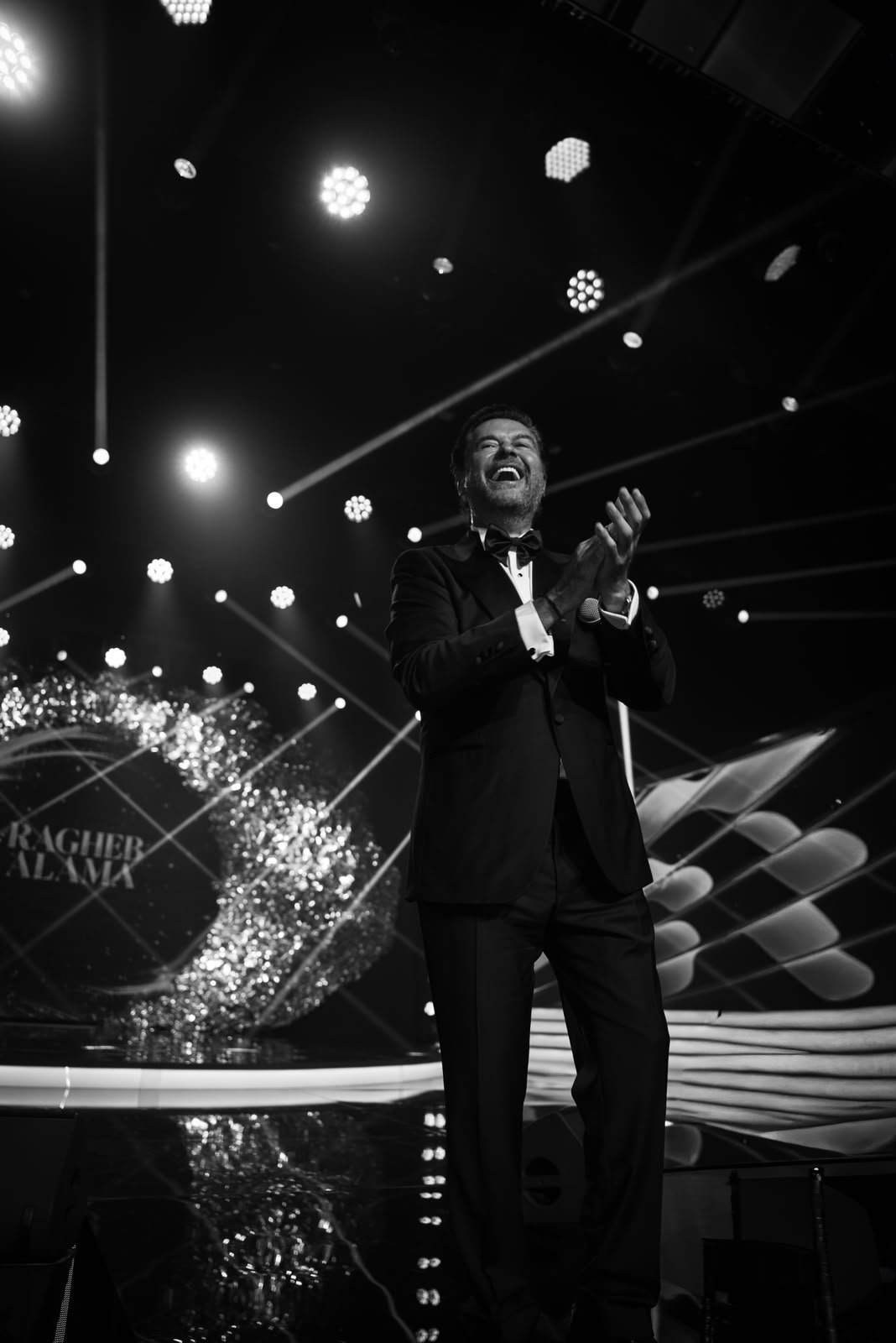 Male singer in a black tuxedo on stage, clapping and smiling under bright spotlights.