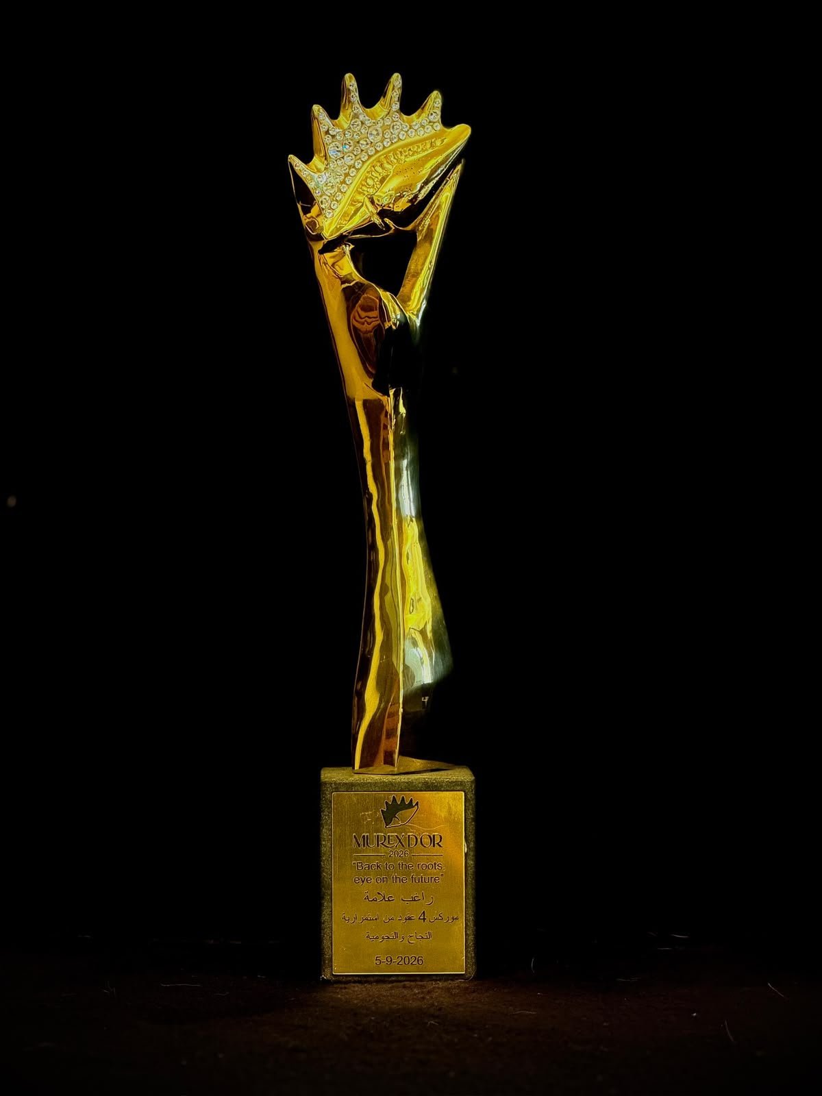 Golden trophy on a black background, with a crowned figure at the top and a plaque on the base.