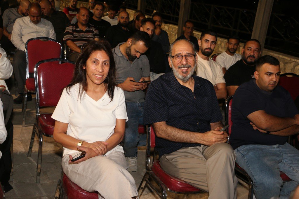 WhatsApp Image 2026-09-07 at 122847 AM - عروبة الاخباري | Oroba News Woman in a white top and beige pants sits smiling in the front row next to a bearded man with glasses at a crowded indoor event.