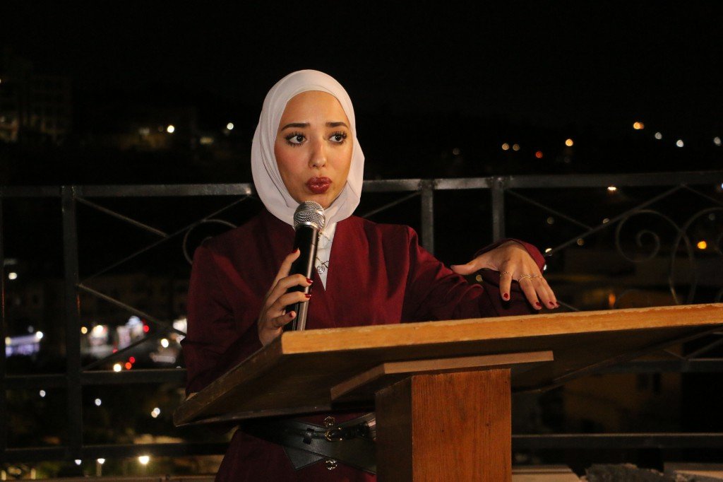 WhatsApp Image 2026-09-07 at 122849 AM - عروبة الاخباري | Oroba News A woman wearing a white hijab speaks into a microphone at a wooden podium during a nighttime outdoor event.
