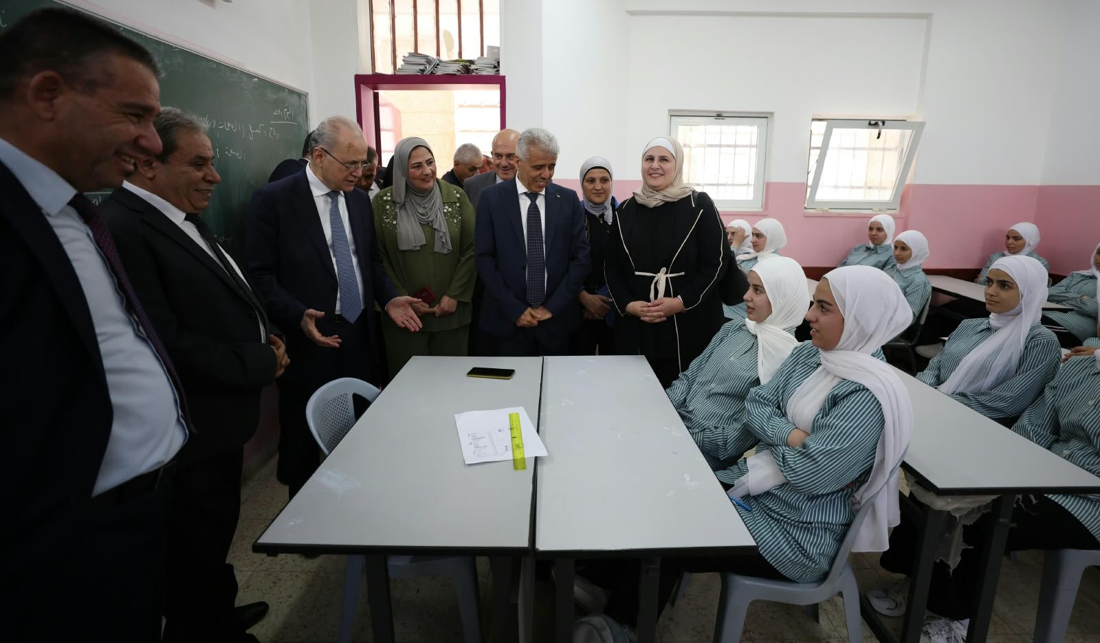 Group of officials visiting a classroom with hijab-wearing students seated at desks near a chalkboard.