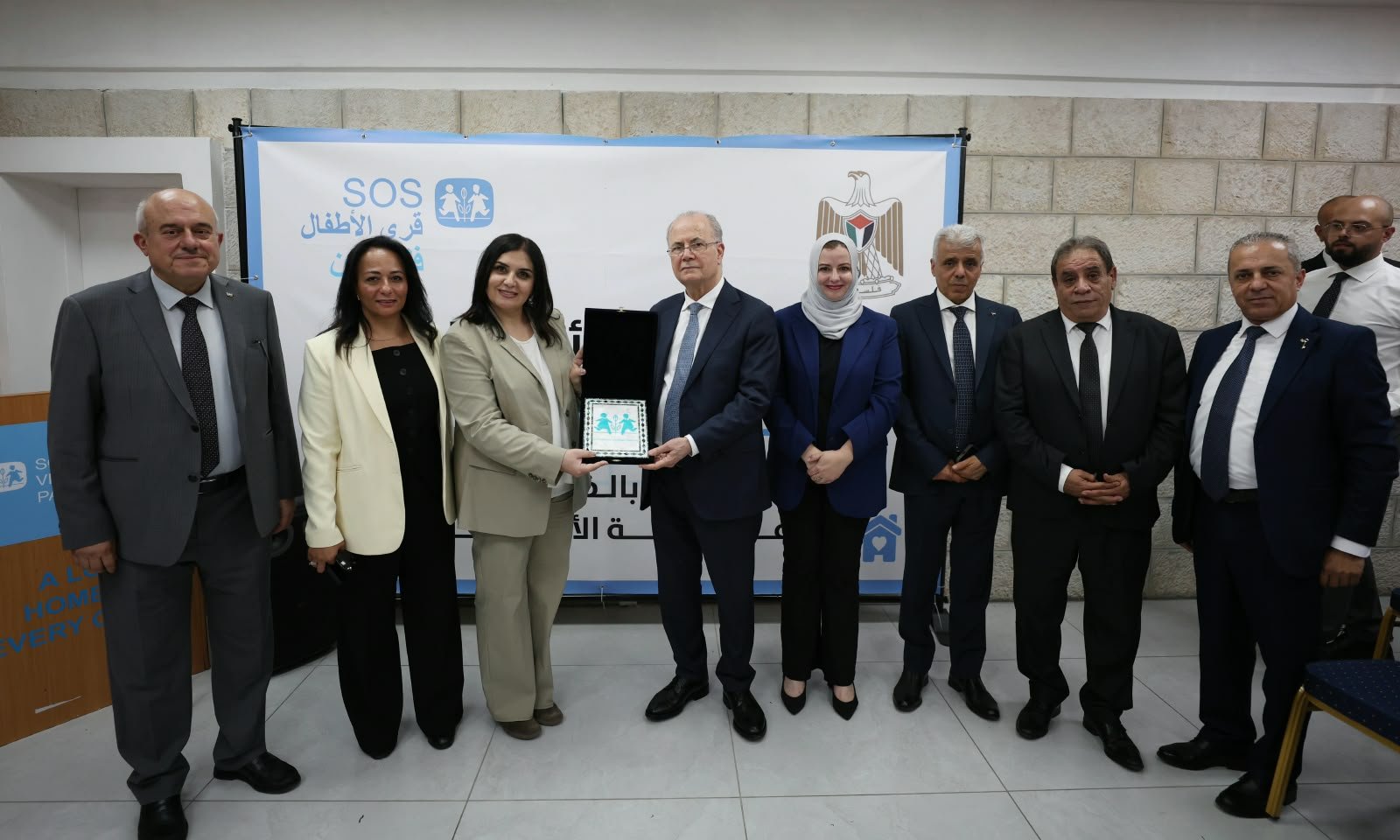 Group of officials in formal attire posing for a ceremony; a man and two women near the center hold a small award plaque in front of a banner.