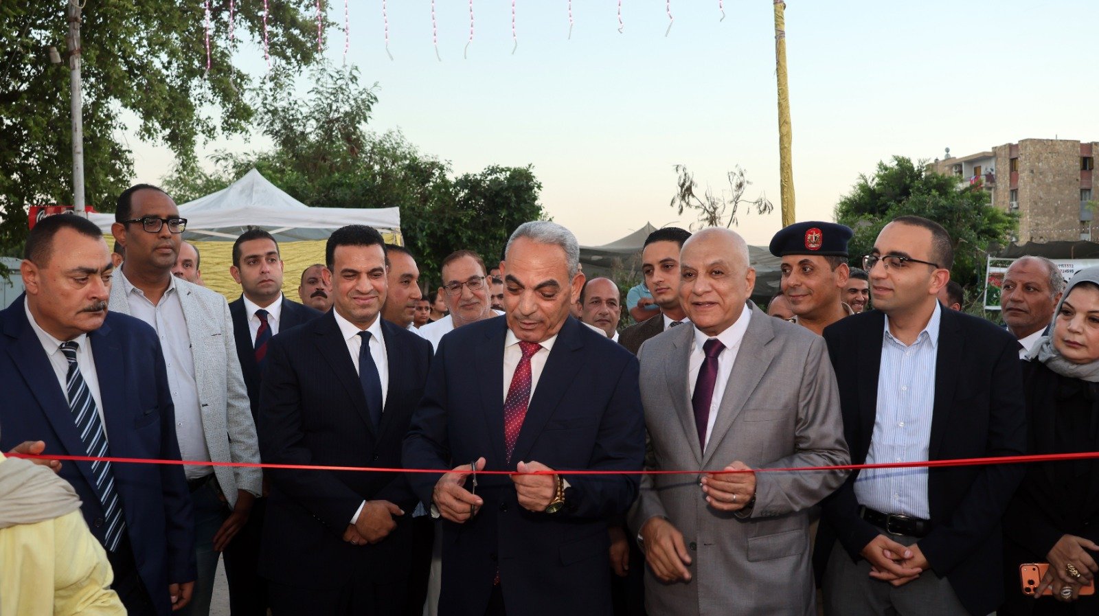 Officials cut a red ribbon at an outdoor ceremony, surrounded by suited guests and onlookers.