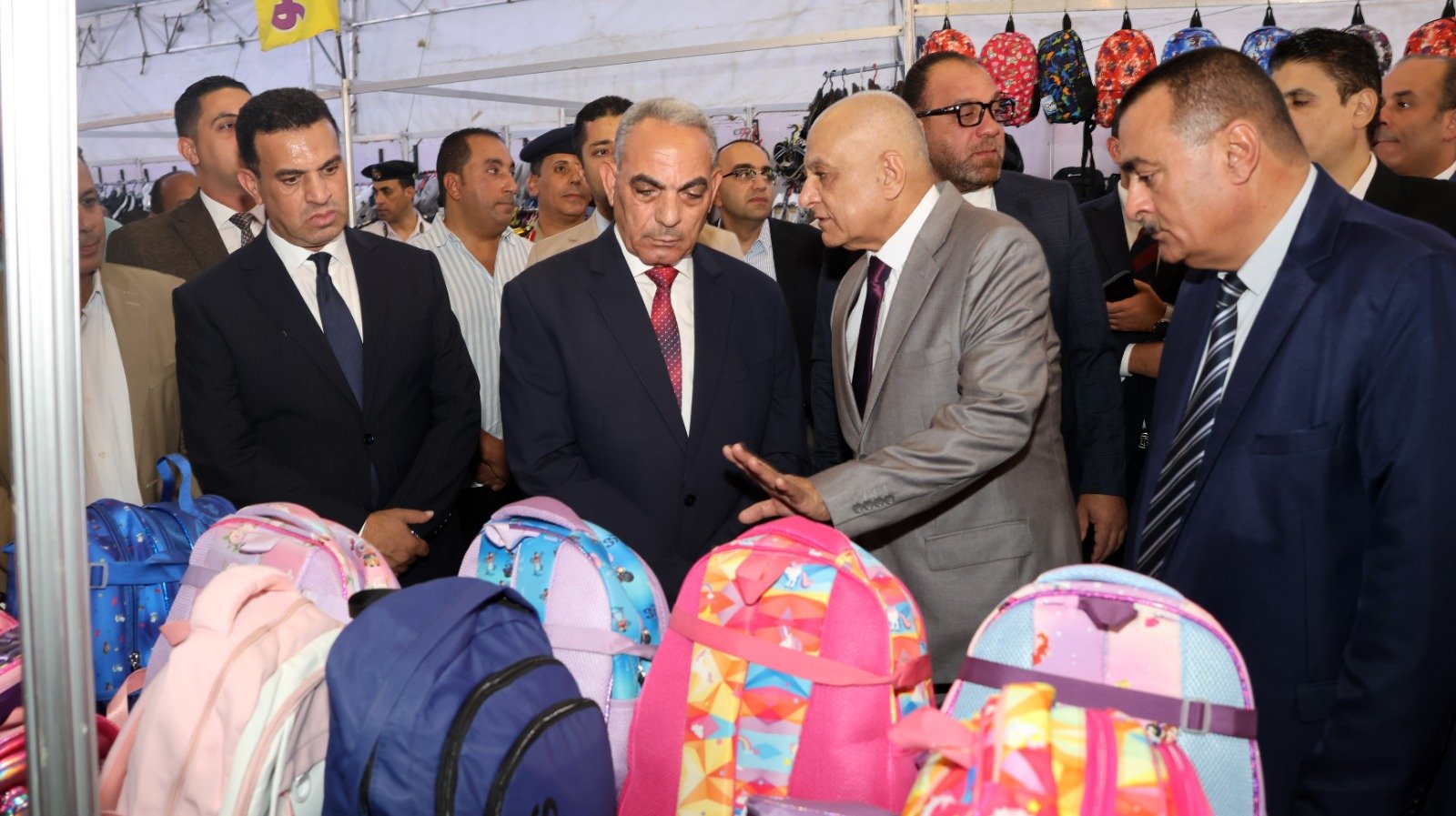 Group of men in suits inspecting colorful backpacks at a market stall or trade show backdrop of hanging bags and backpacks men in formal attire central figure gestures as others listen