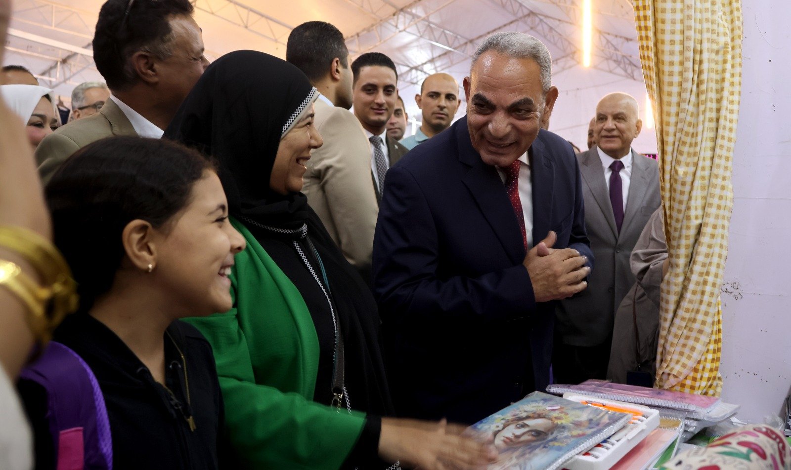 Group of people smiling and talking at a book display, a man in a dark suit greets a woman in a black hijab and green outfit.