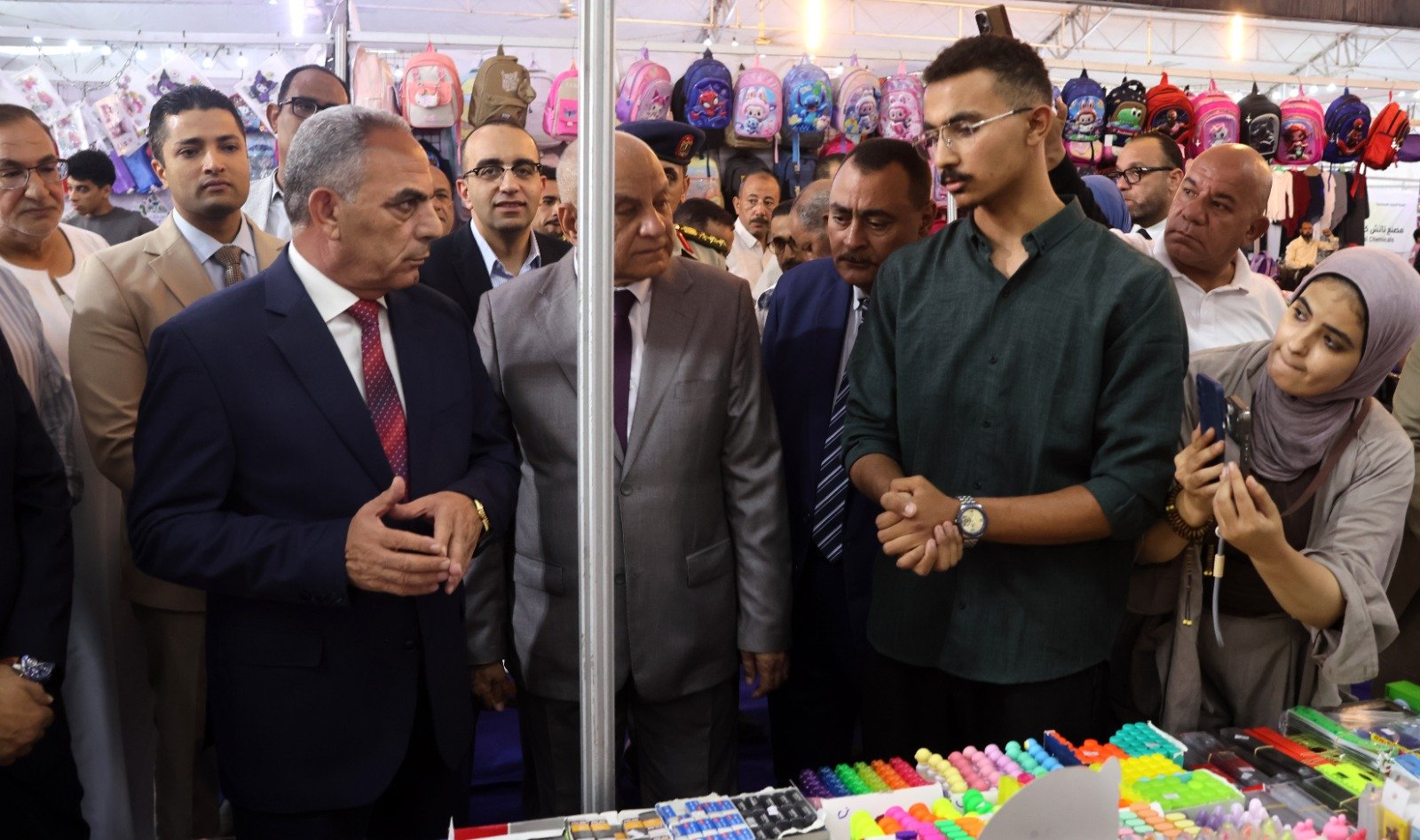 Group of men in suits and a hijab-wearing woman inspect a busy market stall displaying colorful backpacks and stationery items.